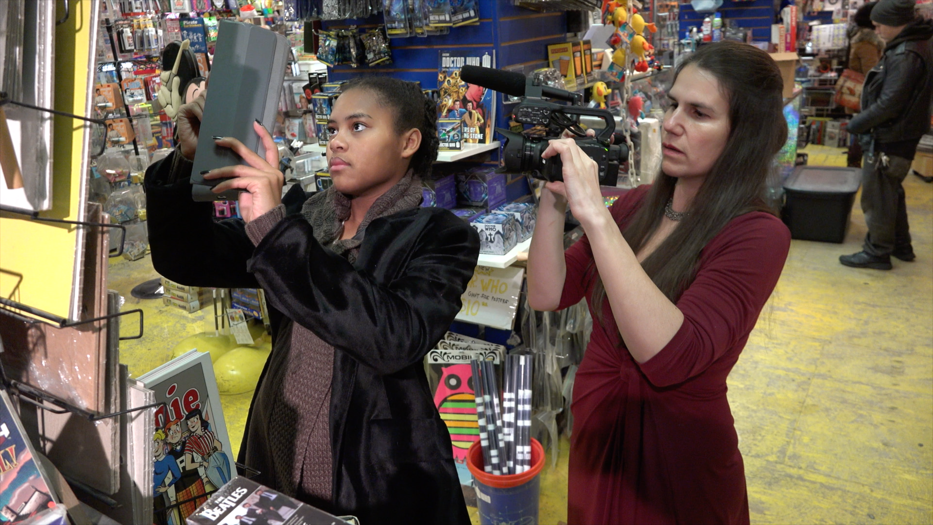 a young women in a comic book shop holds up an Apple ipad. alexander hidalgo is filming the young women with a video camera